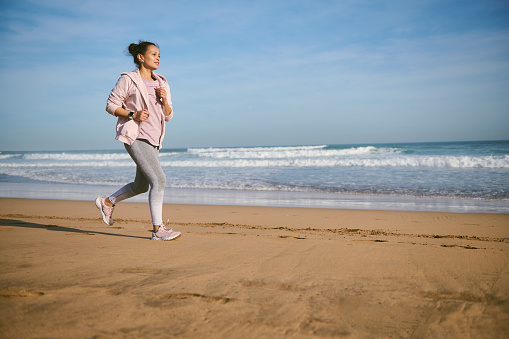 Authentic portrait of a young woman running along a sandy beach in the morning. Happy female athlete in gray leggings and pink t-shirt, enjoying morning jog with Atlantic beach in the background