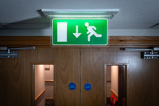 Shallow focus of a generic green, illuminated Fire Escape sign seen next to fire doors in a luxury British hotel. The double doors act as fire doors during an emergency.