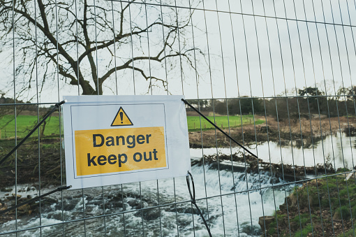 Shallow focus of a Danger Keep Put sign seen next to a barrier which has been erected due to flooding of a nearby English river. The river is seen flowing rapidly after torrential rain.