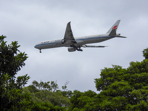 An Air China Boeing B777-39L(ER) plane, registration B-7869, flies over trees in a park as she comes into land from the north at Sydney Kingsford-Smith Airport as flight CA173 from Beijing.  This image was taken from Enmore Park, Marrickville on an overcast afternoon on 24 February 2024.