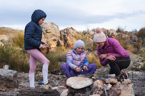 A mother assists her two young girls with their camp fire. They are dressed warm and having fun together outdoors making a fire to heat the water