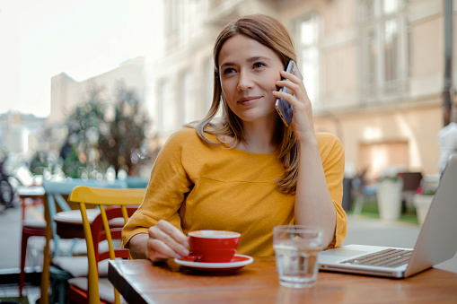 Young charming woman calling with cell telephone while sitting alone in coffee shop during free time, attractive female with cute smile having talking conversation with mobile phone in cafe