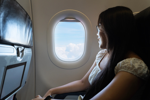 Asian woman with long, dark hair sits in a window seat on an airplane. Looking out the window at the sky, which is filled with fluffy white clouds is smiling and appears to be enjoying the flight.