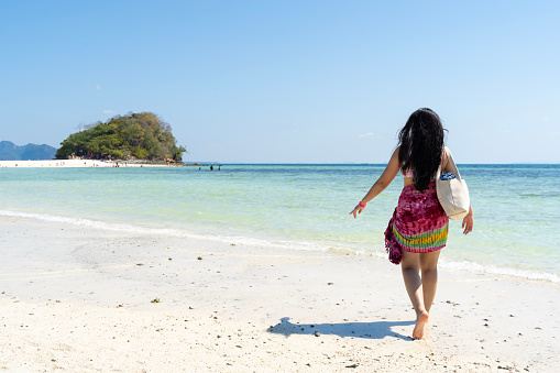 A young woman in a white dress walks along the pristine white sand beach of Railay Beach in Krabi, Thailand. The woman is alone, and she seems to be enjoying the peace and tranquility of the moment.