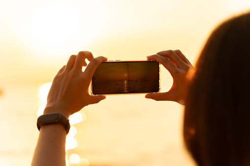 A young woman stands on a beach at sunset, holding her smartphone up to take a picture. The sun is setting in the distance, casting a warm glow over the sky and the water.