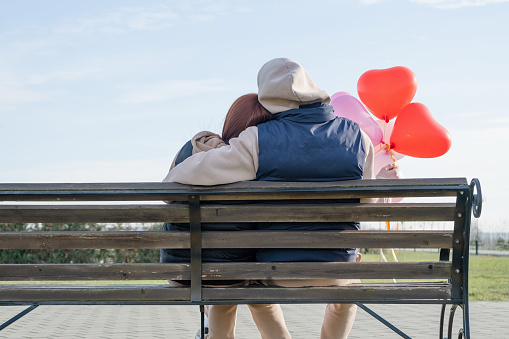 Outdoor photo of happy young woman enjoying date. cheerful romantic couple dating outdoors with balloons, sitting on bench, view from behind