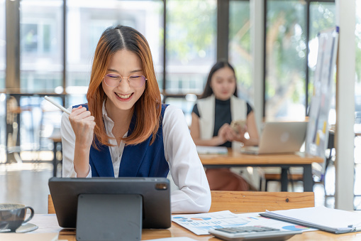 Young gen Z woman working on her project using notebook computer at a co-working space smiling and clenching her fist in excitement while another woman working in the background