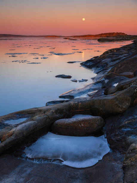Frozen rocks are scattered across the beach as the sun sets in Sillvik, Sweden. The icy formations add a unique and stark element to the coastal landscape.