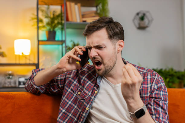 istock Angry young man arguing talking loudly shouting on smartphone quarrel sitting on couch at home 2035305548