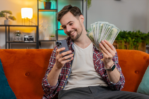 Successful rich man holding smartphone and waving dollar bill money fan sitting on sofa at home. Happy smiling Caucasian guy winning online casino lottery game planning vacation calculating budget