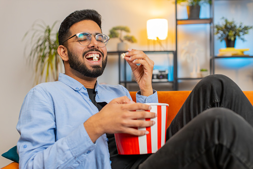 Relaxed Indian young man eating popcorn and watching movie wow expression sitting on sofa in living room at home. Happy Hispanic guy in casual clothes enjoying film during weekend in apartment room.