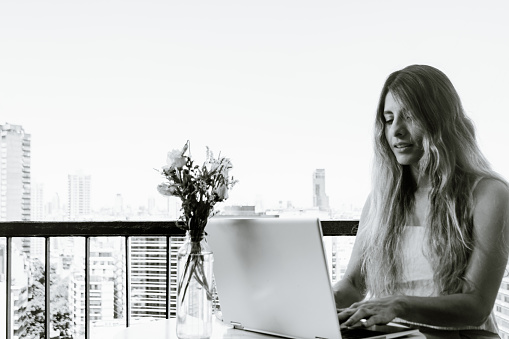 young latin colombian woman on the balcony of her apartment, sitting calmly using her computer, technology concept, copy space. monochrome image