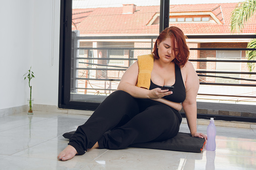 young plus size argentinian hispanic latina white woman with short red hair is sitting checking messages on the phone before exercising indoors, wears black sportswear and yellow small towel.