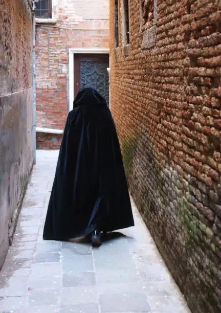 Photo of anonymous hooded stroller with black cloak dress walking through the alleys