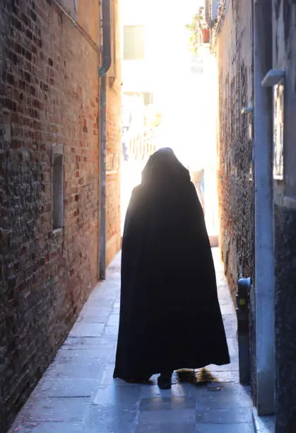 Photo of anonymous hooded stroller with black cloak dress walking through the narrow alleys