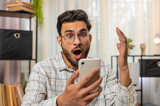 Oh my God, wow. Happy amazed young Hispanic businessman working on smartphone shocked by sudden victory game winning lottery app goal achievement good news. Indian man freelancer guy at home office.