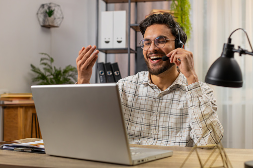 Young Arabian businessman wearing headset, freelance worker, call center or support service operator helpline, talking with client or colleague communication support. Guy sitting at home office desk.