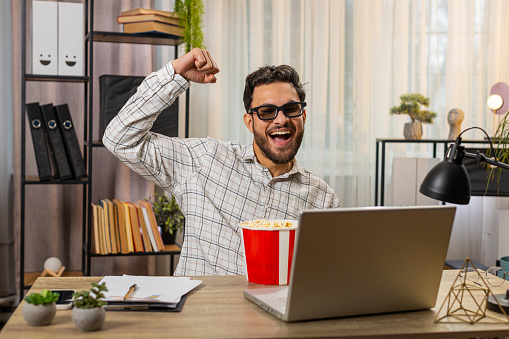 Indian young businessman freelancer taking break from work wearing 3D glasses earing popcorn and watching movie on laptop. Smiling Hispanic man sitting at home office table having snacks during break