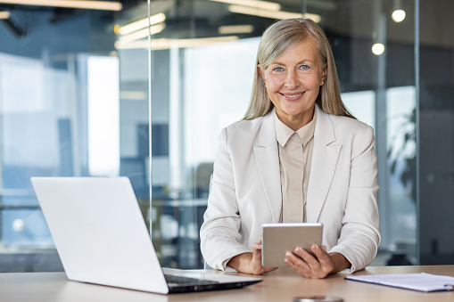 Senior blonde female using digital tablet while sitting by table with portable computer on workspace background. Confident businesswoman using additional modern gadget for increasing productivity.