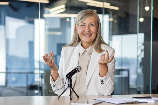 Successful middle aged woman sitting at desk with microphone and smiling while looking at camera. Confident senior businesswoman dressed in formal suit giving interview for press in modern office