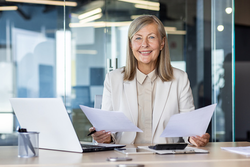 Confident senior businesswoman with documents at office desk, smiling at camera, professional workplace.