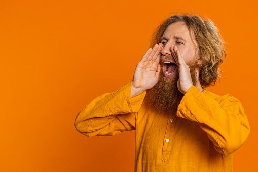 Happy excited Caucasian man yell, shout to camera, promotion advertisement of holidays sale, invitation, greetings, welcome. Excited smiling bearded young guy scream loud isolated on orange background
