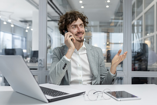 Curly-haired young professional man in a business setting, actively engaged in conversation on a phone call.