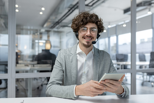 Charismatic young professional with curly hair and headset confidently working in a contemporary office environment, using a tablet.