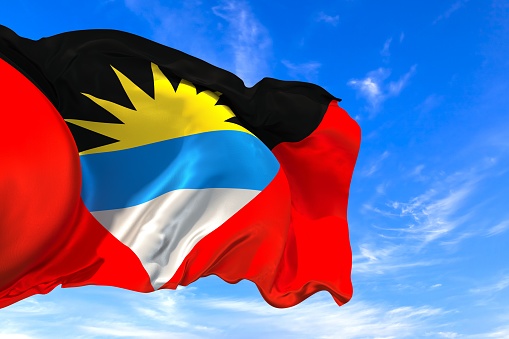 The national flag of Antigua and Barbuda with fabric texture waving in the wind on a blue sky. 3D Illustration