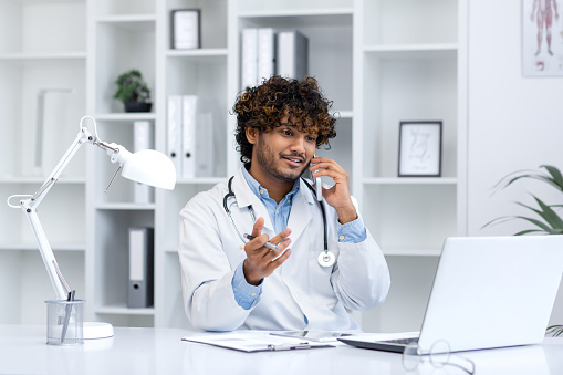 Young successful hispanic doctor inside the clinic in the medical office joyfully communicates talking on the phone, the clinic worker sits at the desk working with a laptop, consults patients.