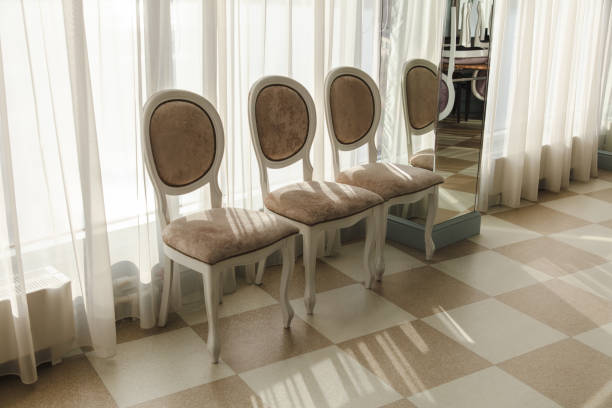 This image highlights a beautifully preserved antique armchair located in the Ambronay town hall, in the mayor's office. The chair, upholstered in rich green velvet, features intricately carved wooden arms and a high back, exuding elegance and historical charm. The backdrop of the image showcases a wall adorned with luxurious red and gold damask wallpaper, complementing the detailed woodwork and stained glass window. Photo taken in Ambronay, in Ain department, in Auvergne-Rhone-Alpes region in France.