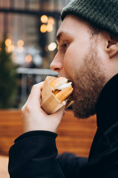 istock A young bearded man eats a burger outside against the backdrop of a cafe. 2035474273