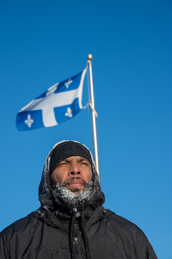 Headshot of black man with frozen beard looking away during winter day with a Quebec flag floating behind on clear sky background