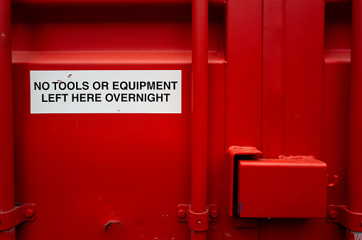 Red painted shipped container with a sticker warning of no tools left in the container. The container is on a building site, used by builders to store tools during the day.