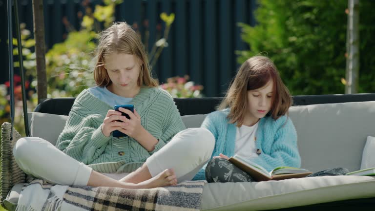 Two friends are relaxing on a garden swing. One girl uses a smartphone, another reads a book