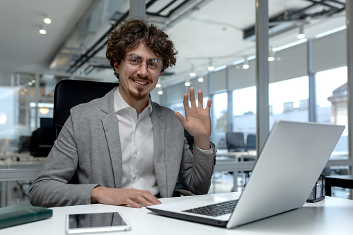 Cheerful young businessman with curly hair waving and smiling in a bright, modern office setting, engaging in a virtual business meeting.