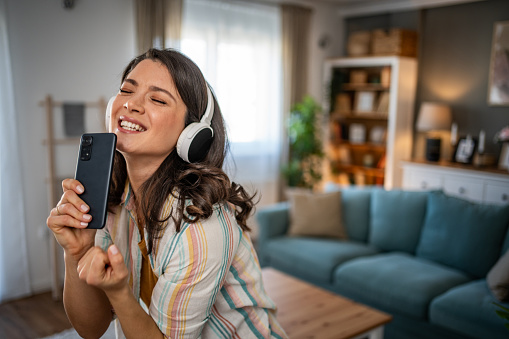 A beautiful young woman is immersed in her world of music as she listens to her favorite songs through wireless headphones connected to her mobile phone. Her face radiates a smile, while her body naturally responds to the rhythm, singing and dancing in the relaxed atmosphere of the living room.