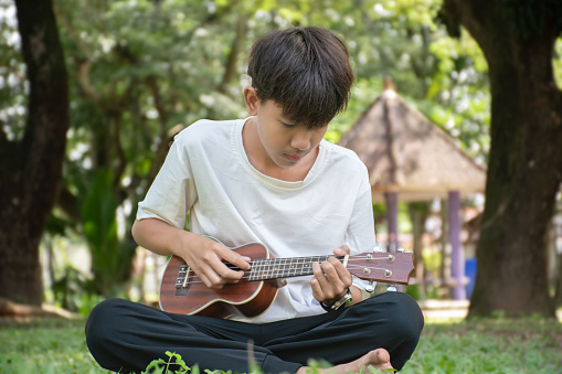 Asian boy spending his freetimes afterschool and on weekend by practising and playing acoustic guitar or ukulele in the backyard of his house.