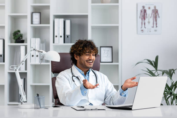 istock Hindu doctor consulting patients remotely, man working inside modern clinic office, using laptop for online video call, talking and smiling friendly to patient 2035569401