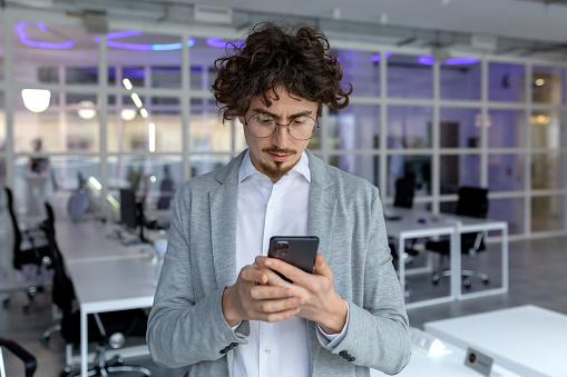 Serious male entrepreneur with curly hair absorbed in reading a text message on his phone at a corporate office.