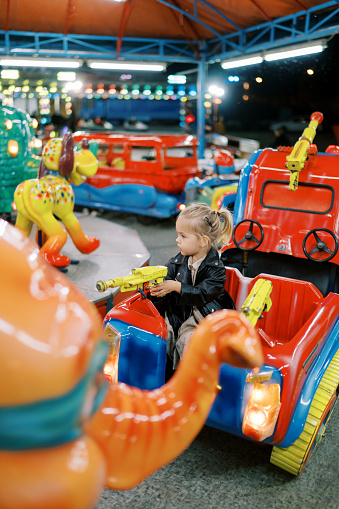 Little girl sits in a toy car on a carousel, holding the pistol steering wheel and looking to the side. High quality photo