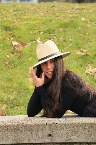 A closeup of a Mexican woman lying on her front on a concrete wall showing off her new white hat. She has long, brown, straight hair, makeup and a black top.