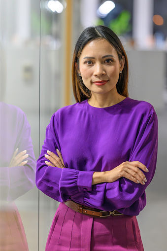 Well dressed mid adult asian woman standing with arms crossed in business office and looking at camera portrait