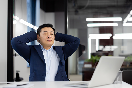 Asian businessman in blue suit stretching while taking a break from work in a bright, contemporary office setting.