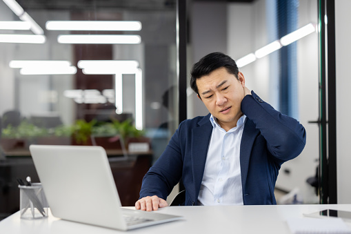 Overworked overtired asian man in business suit at workplace inside office, businessman has severe pain in neck, massaging muscle , long time sitting work with laptop, daily business of financier.