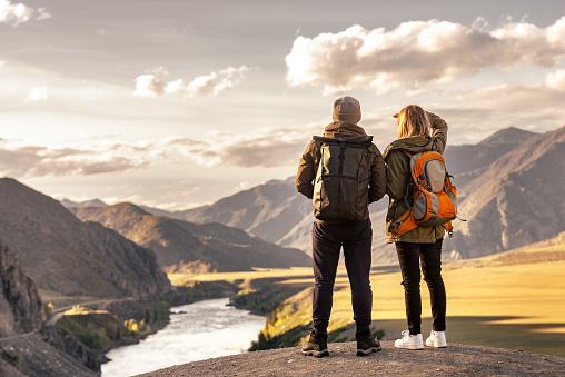 Young couple of tourists is standing with backpacks in mountains and enjoys nice view at sunset and big river