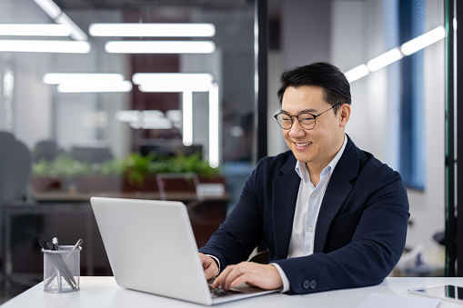 Asian man in business suit and glasses working with laptop inside office, mature businessman typing on keyboard, preparing financial report, male company employee at workplace.