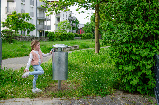 Girl throws trash into a street trash can. Trash can is metal, gray. Child of five with a long blonde braid. She is wearing a cap, pink vest and blue jeans. There's greenery all around.
