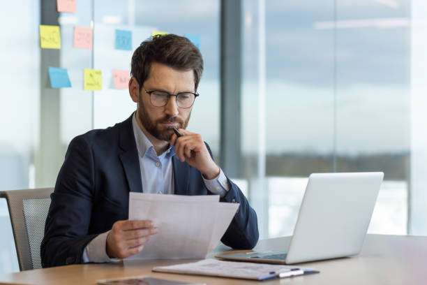istock Serious thinking businessman behind paper work, mature experienced boss working with reports and financial documents, financier inside office with laptop, reading contract, thoughtfully checking 2035817482