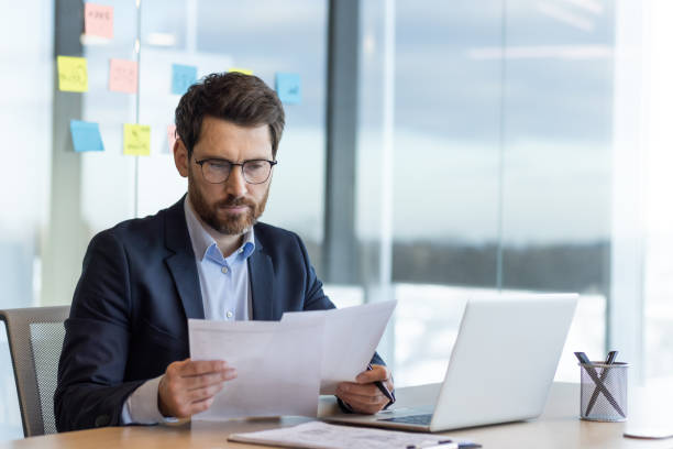 istock Serious thinking businessman behind paper work, mature experienced boss working with reports and financial documents, financier inside office with laptop, reading contract, thoughtfully checking 2035818523